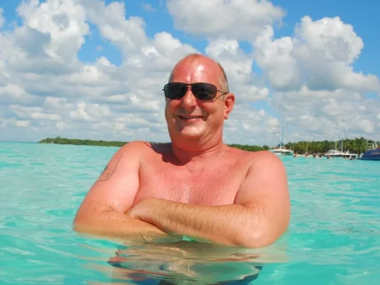 Rainer, sonriendo en el agua turquesa, con gafas de sol y paisaje tropical al fondo.