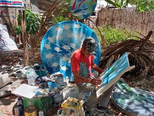 Artista en un estudio al aire libre, trabajando en una pintura con motivos marítimos, rodeado de diferentes botes de pintura y otras obras de arte.