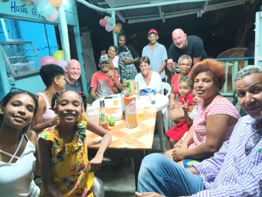 Grupo de personas de diferentes edades, incluidos niños, adultos y personas mayores, sentados y de pie alrededor de una mesa en un evento festivo al aire libre con globos y bebidas.