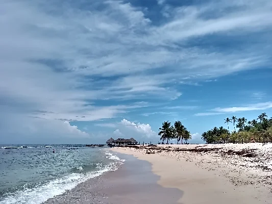 Vista de la playa desde Playa Hemingway en Juan Dolio con cielo despejado, suaves olas, palmeras y un edificio tradicional en el horizonte.